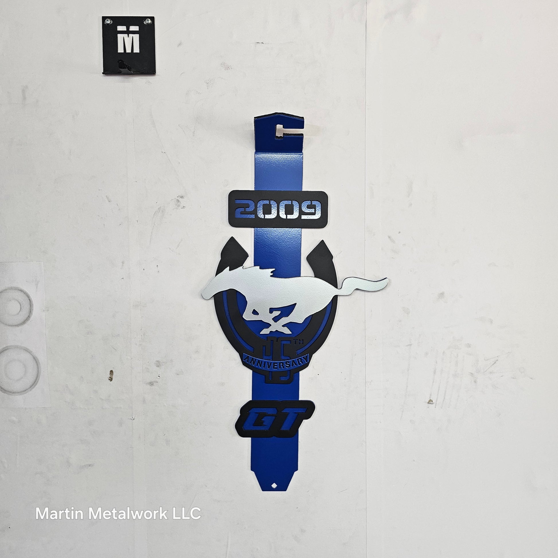 Metallic blue and black car hood ornament on a textured white surface with 'Martin Metalwork LLC' branding.