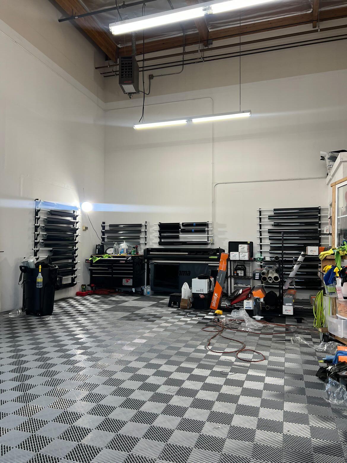 Comprehensive view of a modern vehicle wrap shop with organized wall-mounted vinyl racks, advanced printing equipment, and a striking checkerboard floor. The spacious interior is well-lit, highlighting the clean and professional environment designed for vehicle customization.