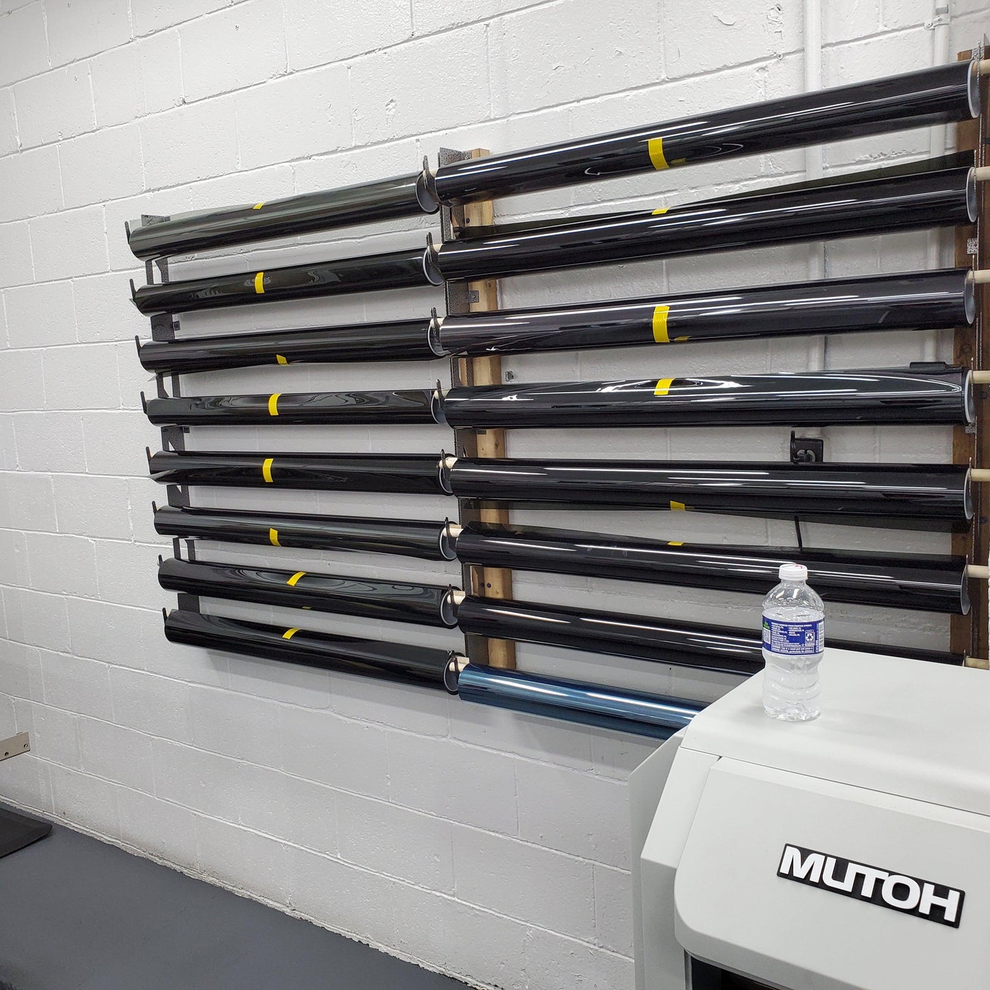 "Organized wall-mounted vinyl wrap rack in a professional print shop, featuring multiple levels of labeled tubes with black vinyl rolls, each marked with yellow stickers. This storage solution is set against a grey cinder block wall, optimizing the use of space in a busy working area."