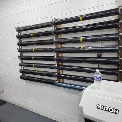 "Organized wall-mounted vinyl wrap rack in a professional print shop, featuring multiple levels of labeled tubes with black vinyl rolls, each marked with yellow stickers. This storage solution is set against a grey cinder block wall, optimizing the use of space in a busy working area."