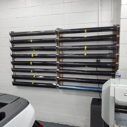 Industrial vinyl wrap and tint storage rack mounted on a white brick wall, featuring multiple horizontal rods with yellow labels for organization, next to office equipment including a large format printer and a water bottle for scale.