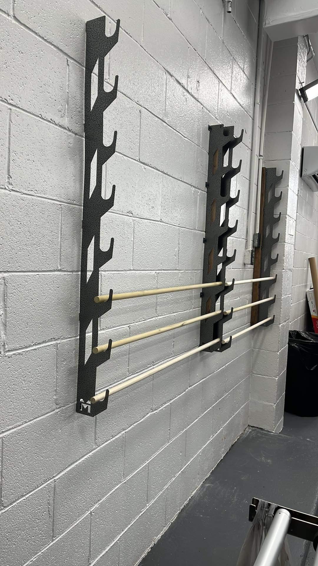 Wall-mounted storage system for vinyl wraps and tints, featuring two black metal brackets with multiple slots for holding poles, which are displayed with three poles installed. The brackets are affixed to a white cinder block wall in a workspace, with the company's logo visible on the bottom bracket.