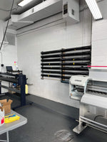 Professional vinyl wrap and tint rack system mounted on a clean white cinder block wall in a well-lit workshop, flanked by large format printers. The rack holds multiple rolls of tint, neatly organized for accessibility and efficiency in the workspace.