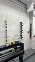 Modern workshop setup with a wall-mounted, bracket-based vinyl wrap and tint storage system next to a digital large format printer. Two vertical racks with cut-out slots are holding three horizontal poles against a white painted cinder block wall, showing a clean and organized work environment.