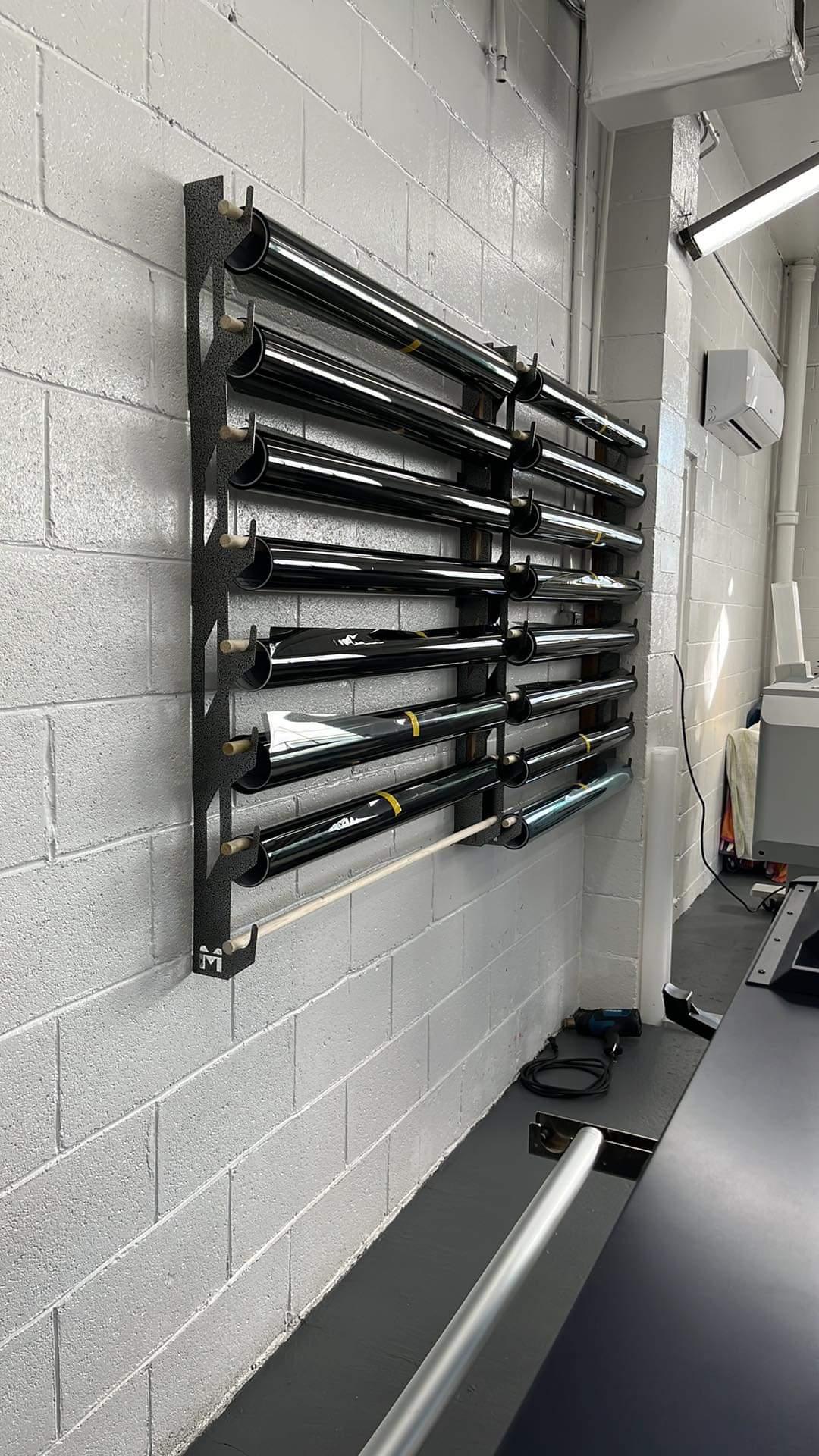 Professional vinyl wrap and tint rack system mounted on a clean white cinder block wall in a well-lit workshop, flanked by large format printers. The rack holds multiple rolls of tint, neatly organized for accessibility and efficiency in the workspace.