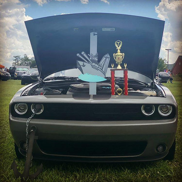 "grey dodge challenger with an open hood displaying a custom hood prop featuring a boat and anchor design, complemented by a trophy placed in front of the engine bay, at an outdoor car show."